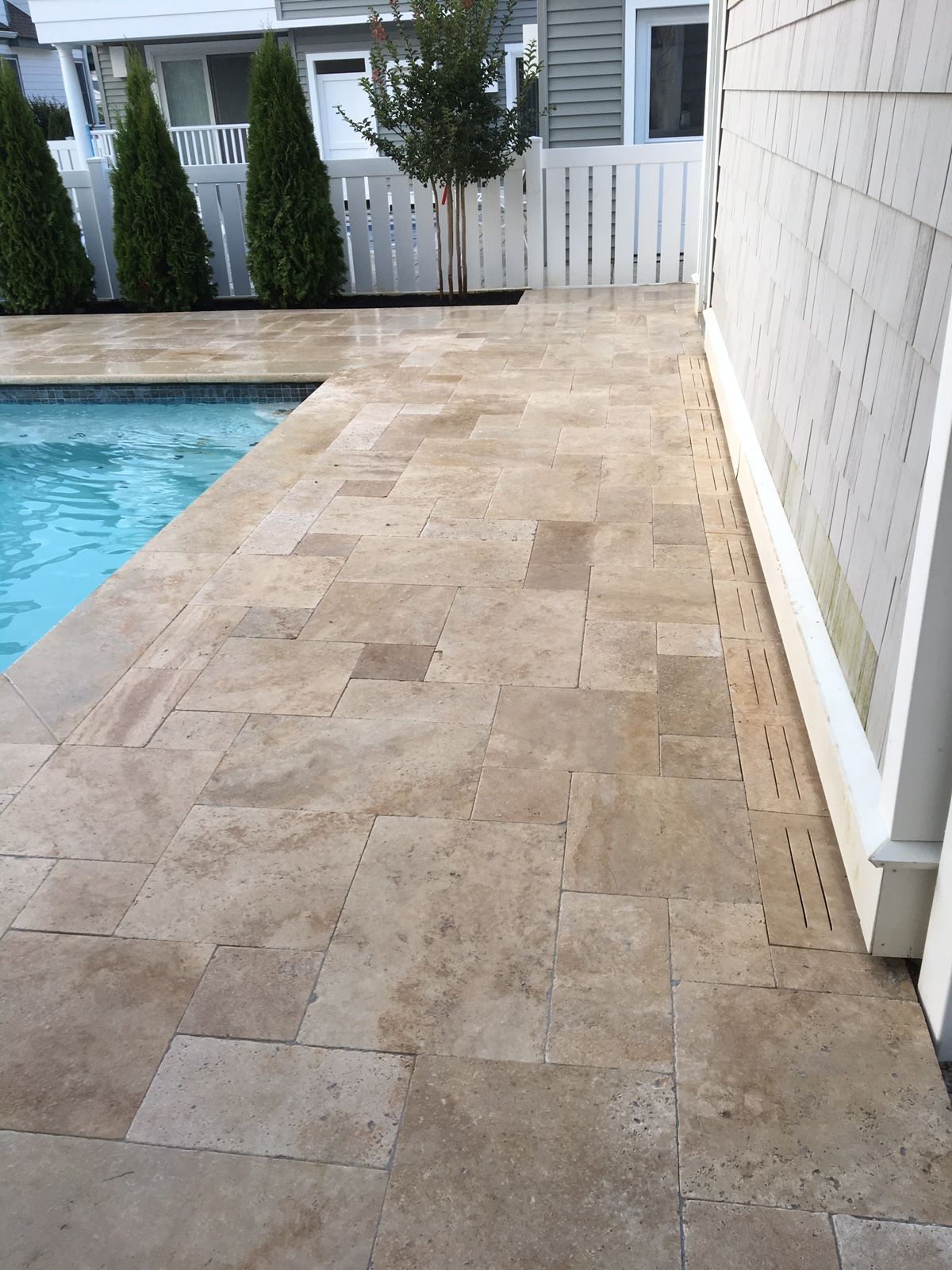 Poolside patio with travertine tiles, bordering a pool, a house, and a white fence.
