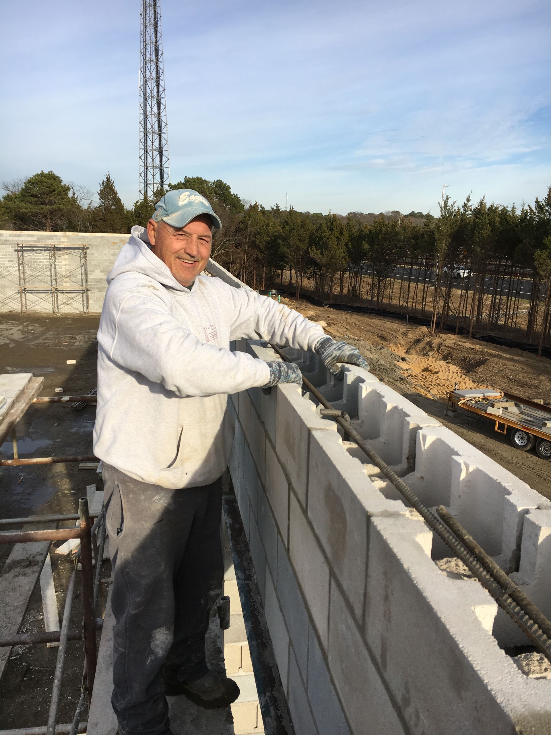Construction worker, in gloves, standing next to a cinder block wall, with rebar, on a sunny day.