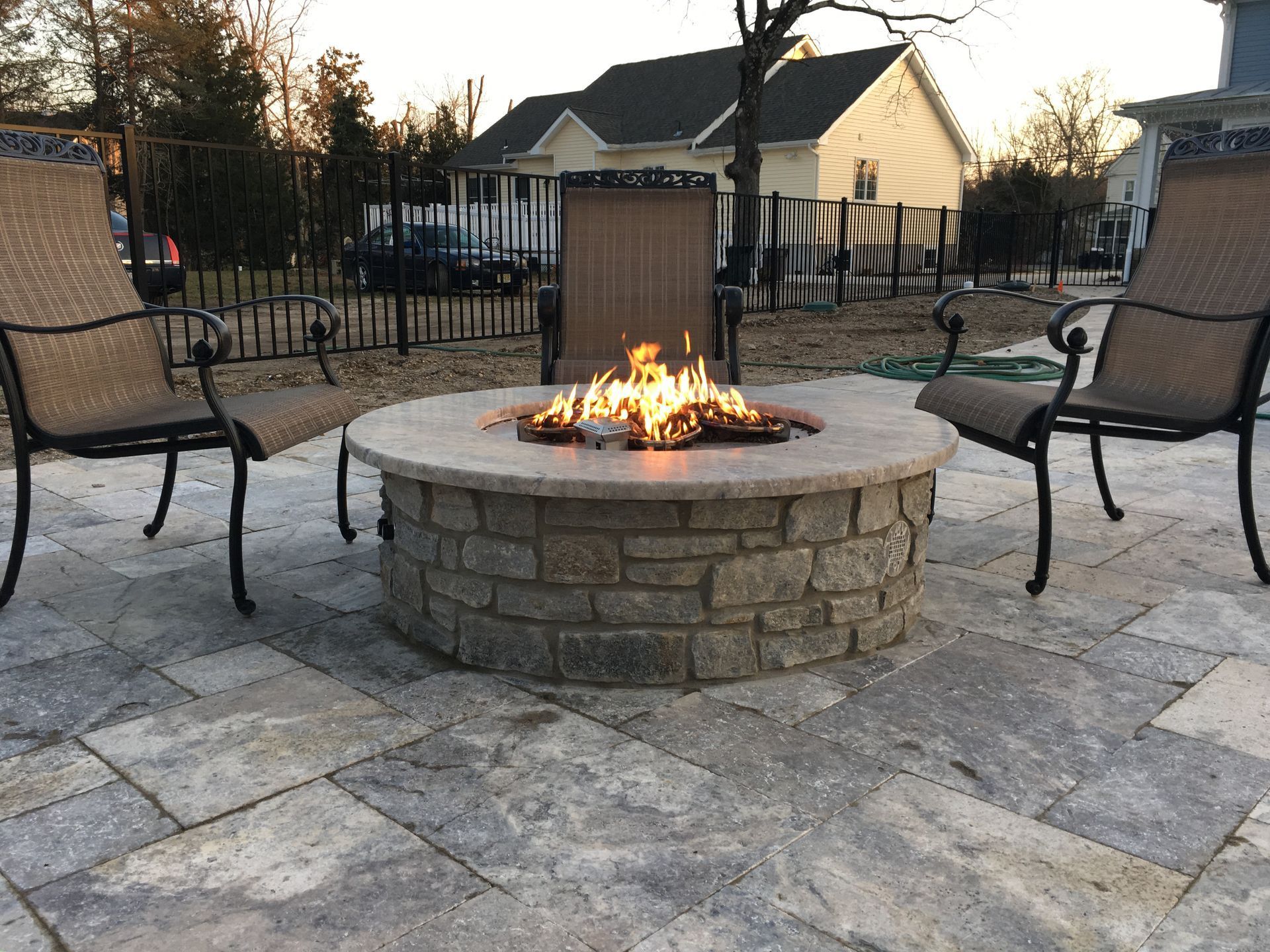 Round stone fire pit with flames, three chairs on stone patio, residential backyard setting.