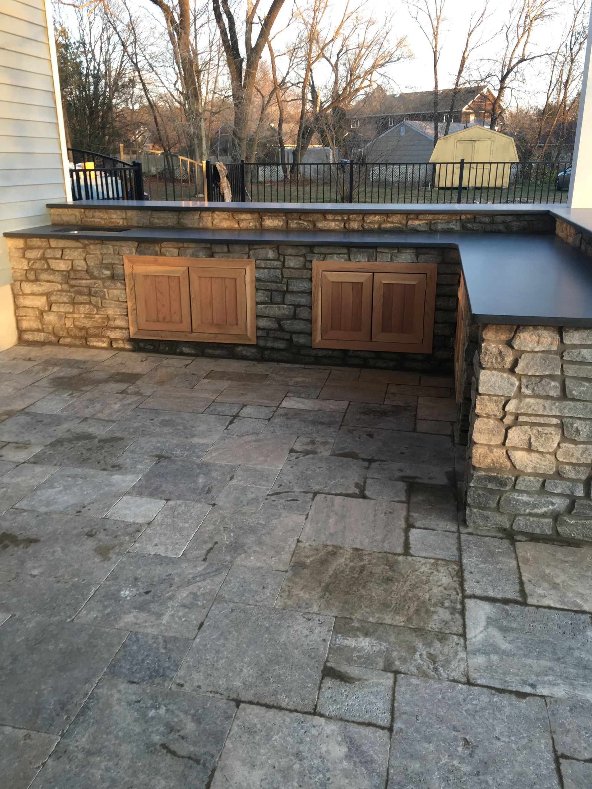 Outdoor stone kitchen with gray countertops and wooden cabinet doors on a flagstone patio.