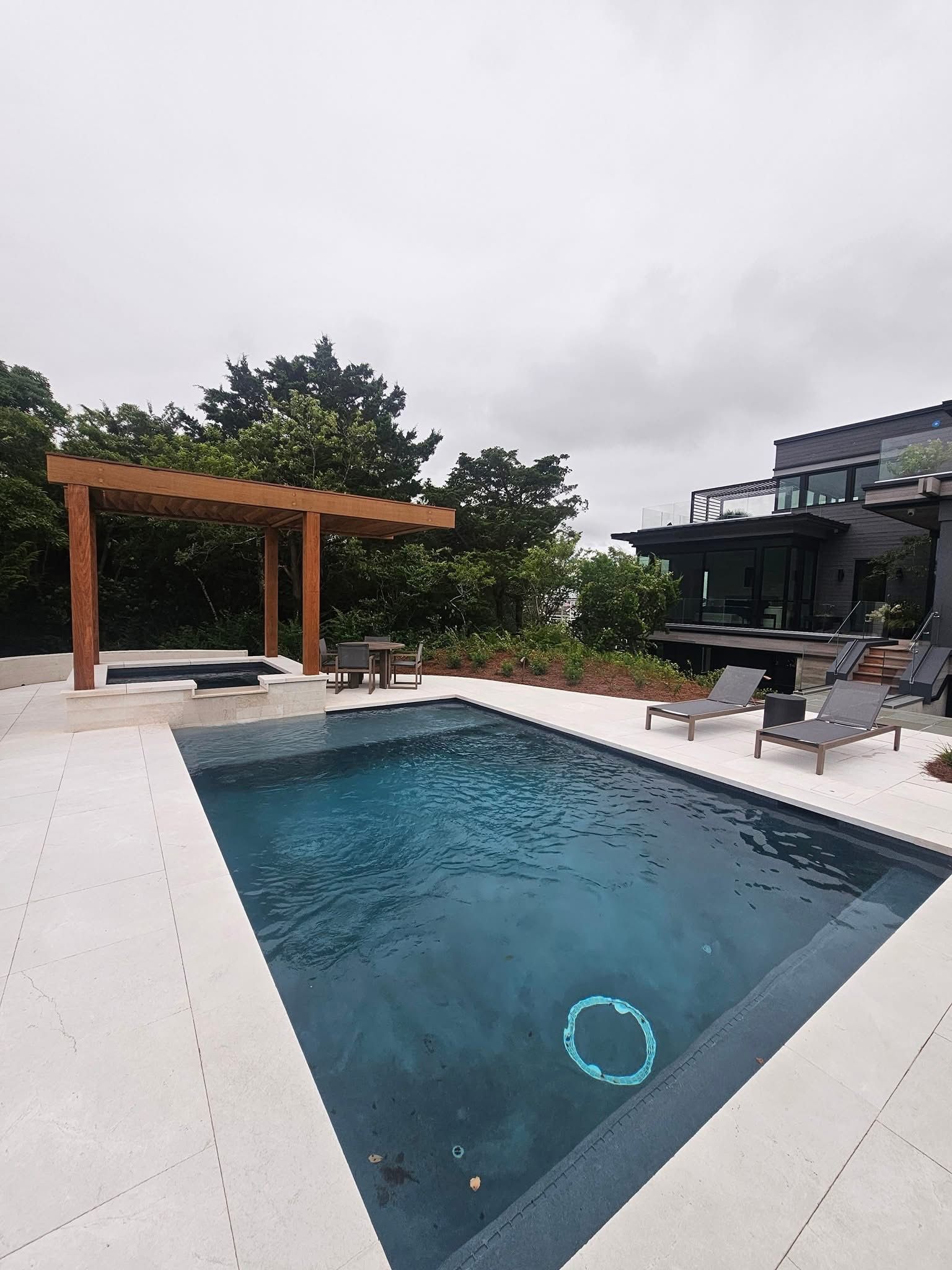 Outdoor swimming pool with pergola, lounge chairs, and modern house under overcast sky.