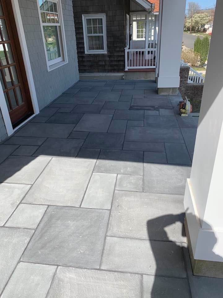 Gray stone patio with rectangular and square pavers in front of a house.