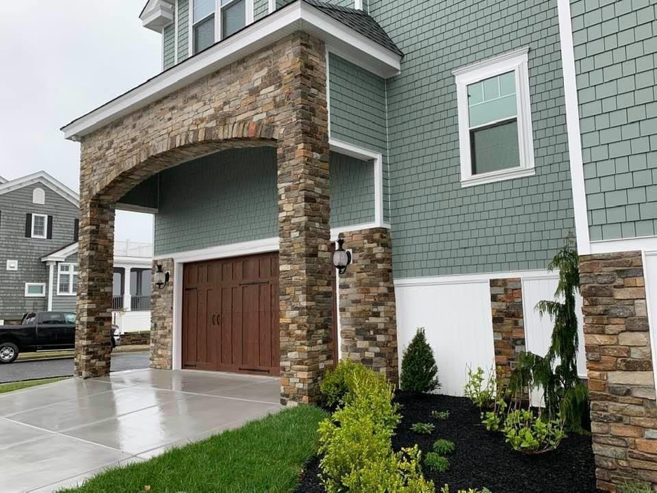 Stone-columned garage entrance with brown door. Teal siding, white trim, and a small garden.