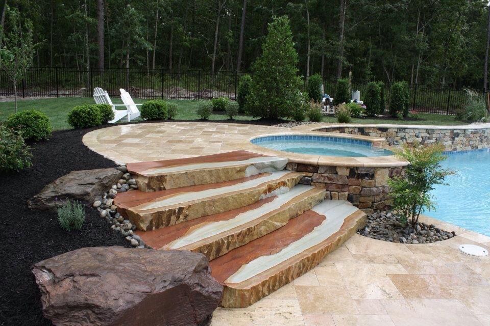 Stone steps lead to a pool and hot tub, surrounded by tan patio and black mulch, trees in background.