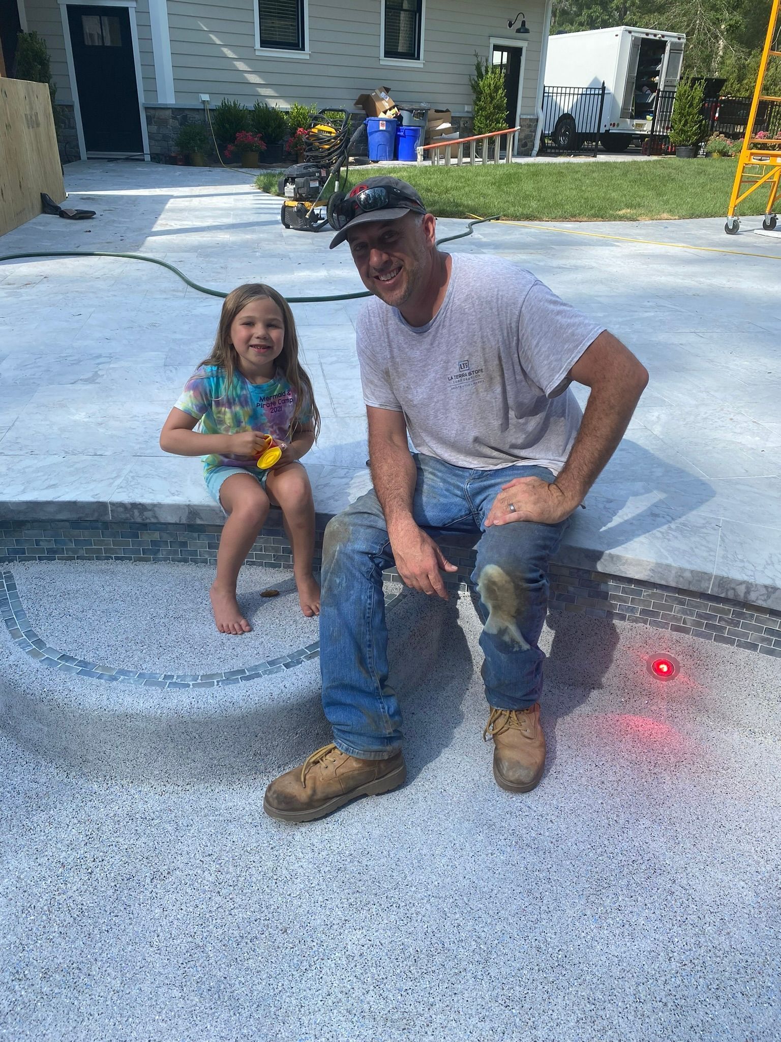Man and child sit on a light-colored surface; building in background. Man smiles, wearing boots, jeans. Child smiles, wearing shorts.