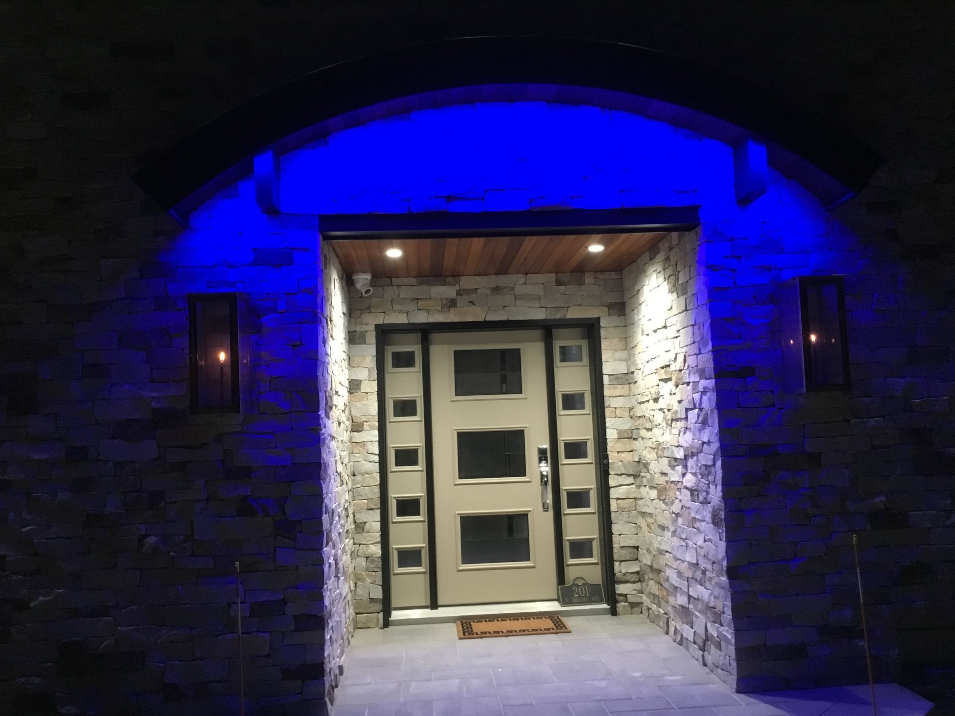 A modern house entrance at night, blue and white lights illuminate stone walls and front door.