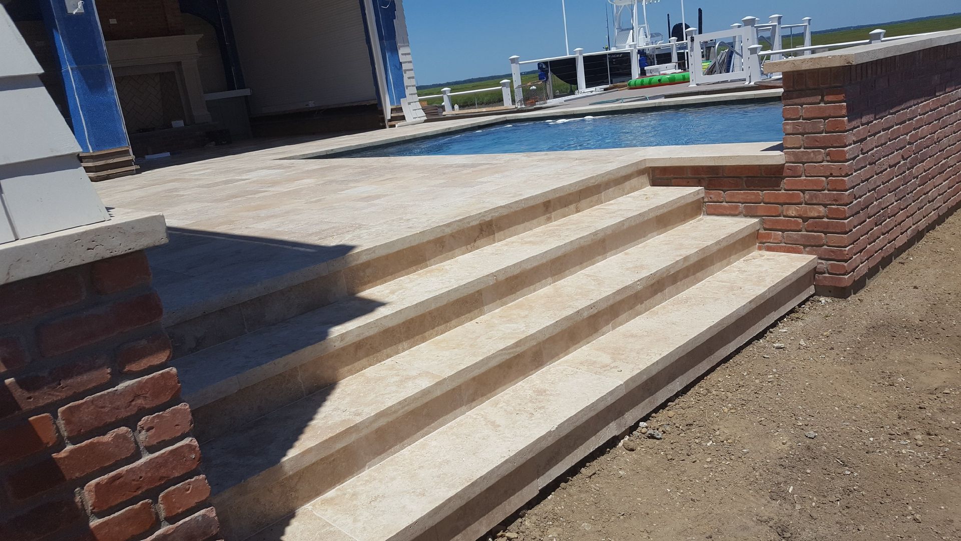 Brick steps lead to a pool with a brick wall. Light-colored patio and blue building in the background.