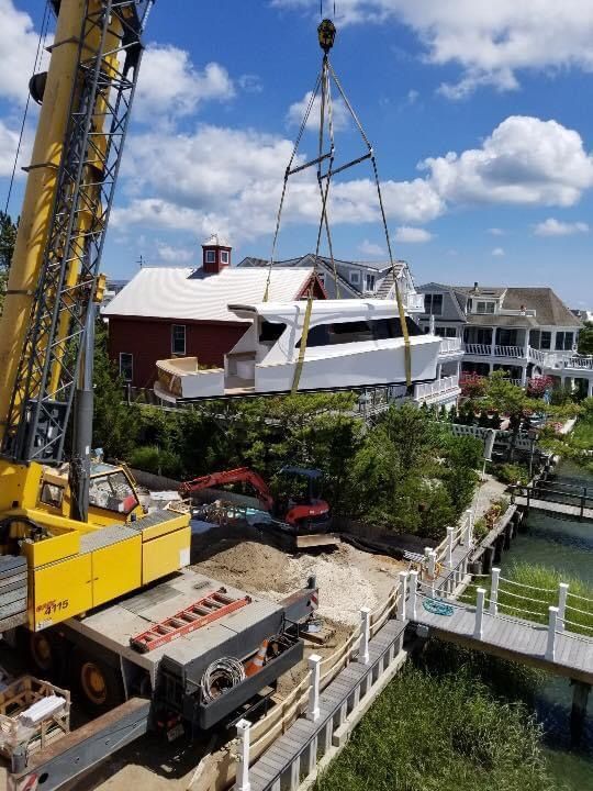 A large yellow crane lifting a white boat over a construction site near a canal, with houses and a red barn in the background.