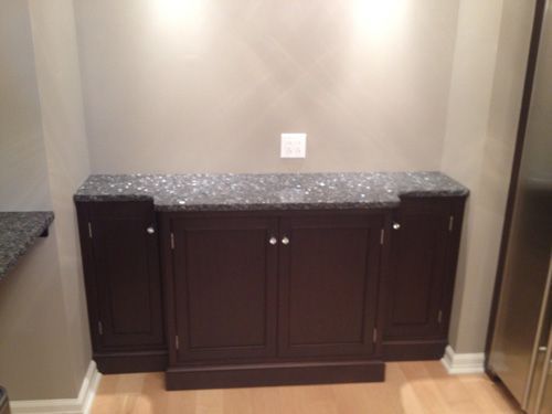 Dark brown cabinet with granite countertop, set in a recessed wall, light wood floor, and silver refrigerator visible.