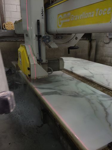 Marble slab being cut by a large machine with laser guides. Yellow saw head, light grey slab.