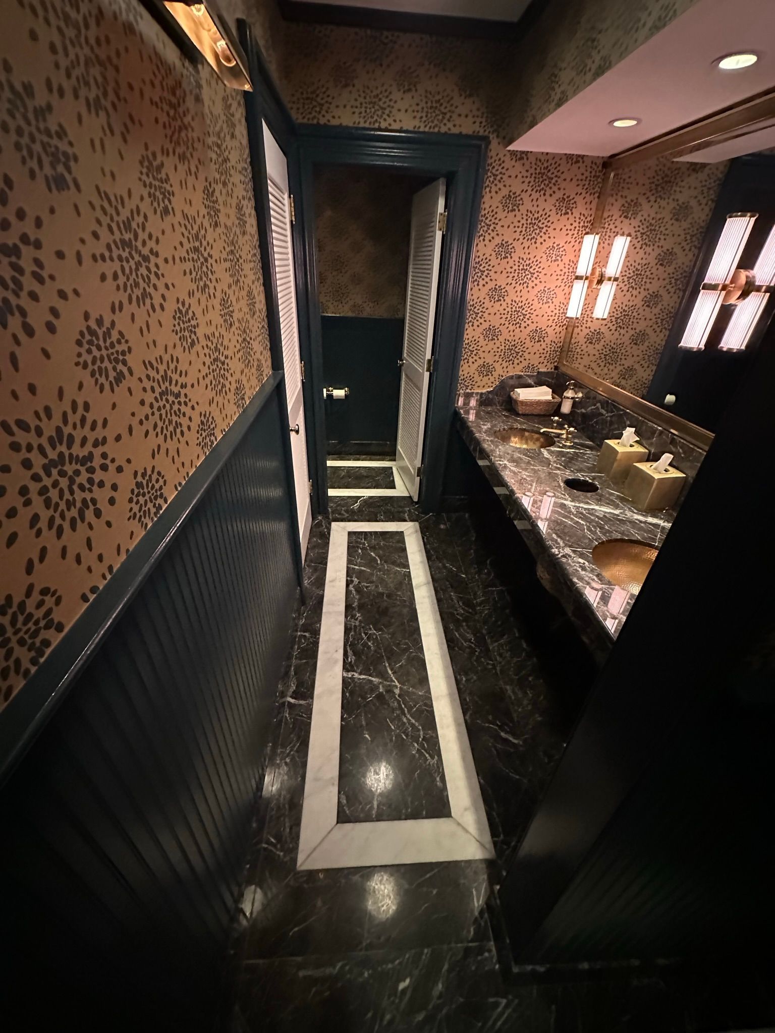 Narrow bathroom with floral wallpaper, black trim, and marble floor with white borders.