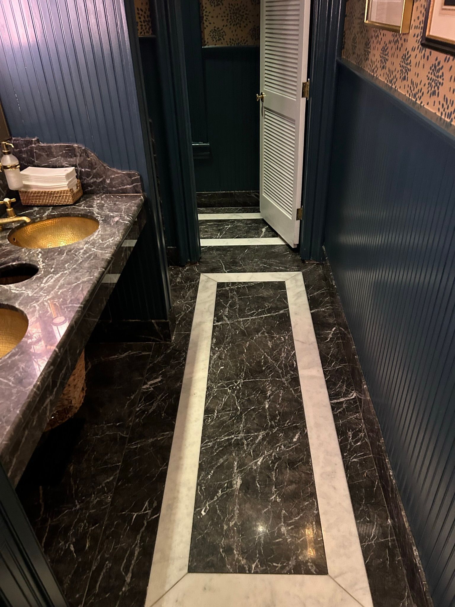 Bathroom with marble floors, gold sinks, blue walls, and a white door leading to a stall.