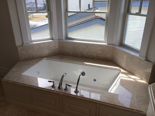 Built-in bathtub with beige marble surround under a bay window; chrome faucet fixtures.