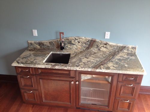 A bar area with granite countertop, sink, cabinets, and a mini-fridge against a blue wall.