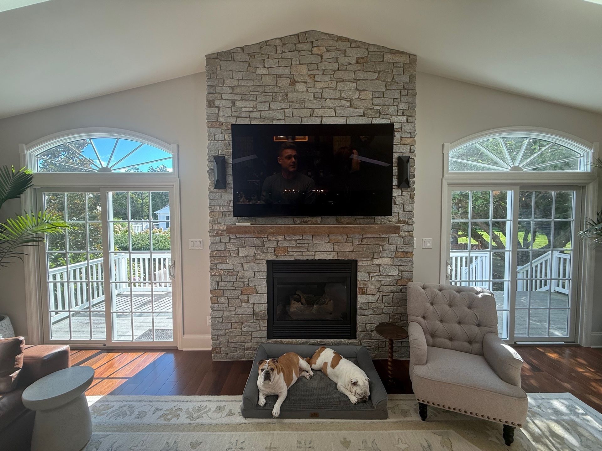 Living room with stone fireplace, TV, dogs on bed, arched windows, and a chair.
