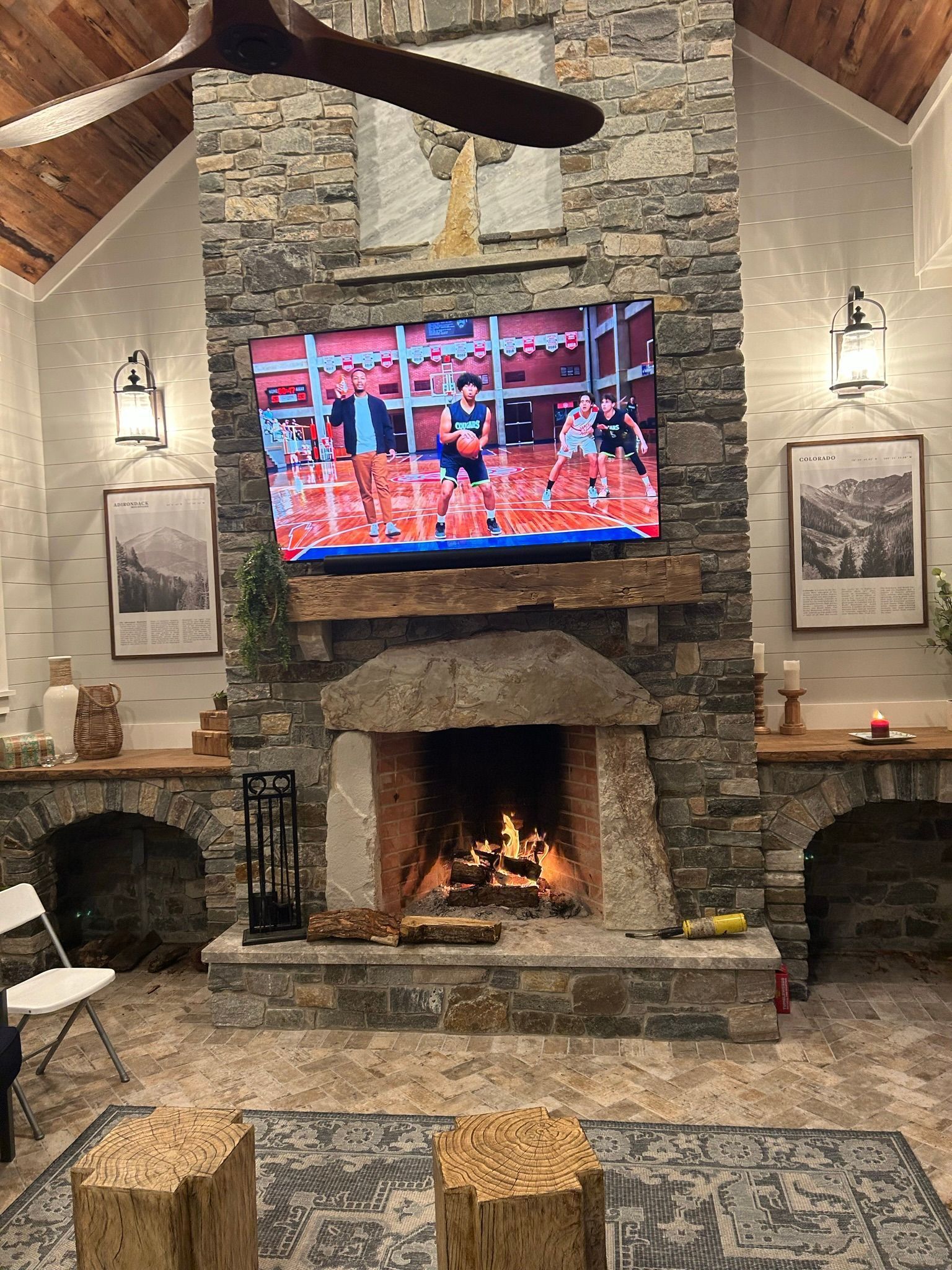 Cozy living room with stone fireplace, TV, and lit fire. Two wooden stools sit in front of the fireplace, a white chair to the left.