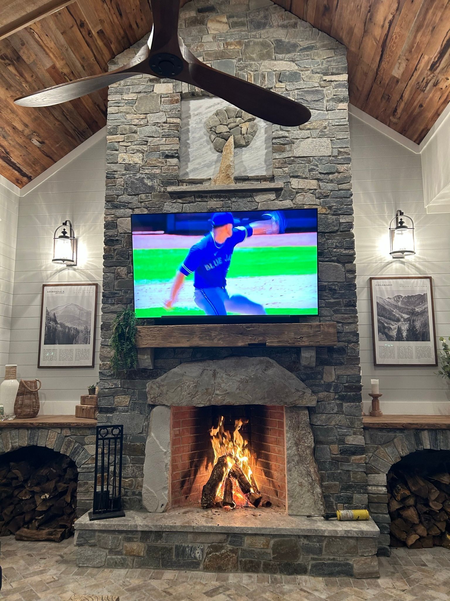 Cozy living room with stone fireplace, TV, and lit fire; wood ceiling and walls, watching a baseball game.