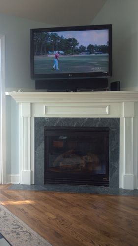 TV above fireplace displaying golf. Fireplace has green tile and white trim. Wooden floor.