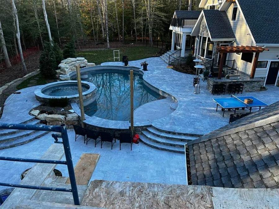 Backyard with pool, hot tub, patio, and house. Blue pool water, gray stone patio, green lawn, trees.
