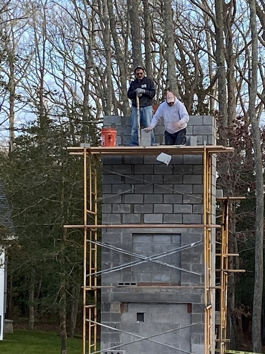Two people on scaffolding building a tall gray cinder block structure outdoors, trees in background.