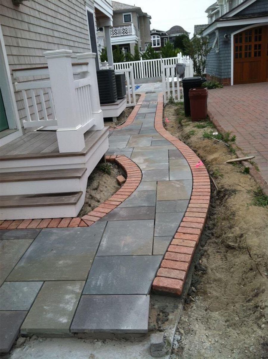 Stone walkway with a brick border curves along a house and to the right; sand present.