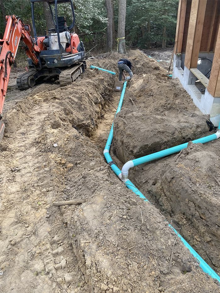 Construction worker installing blue PVC pipes in a trench next to a building. An excavator is nearby.