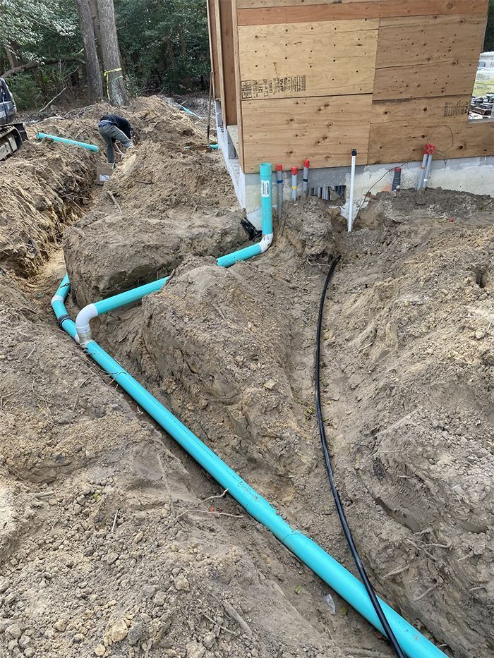 Construction site: trench with blue PVC pipes, electrical conduit, and a worker by unfinished building.