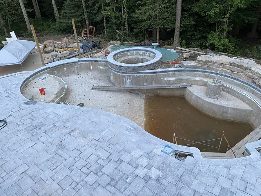 Construction site: unfinished freeform pool and jacuzzi, surrounded by stonework, with wooded backdrop.
