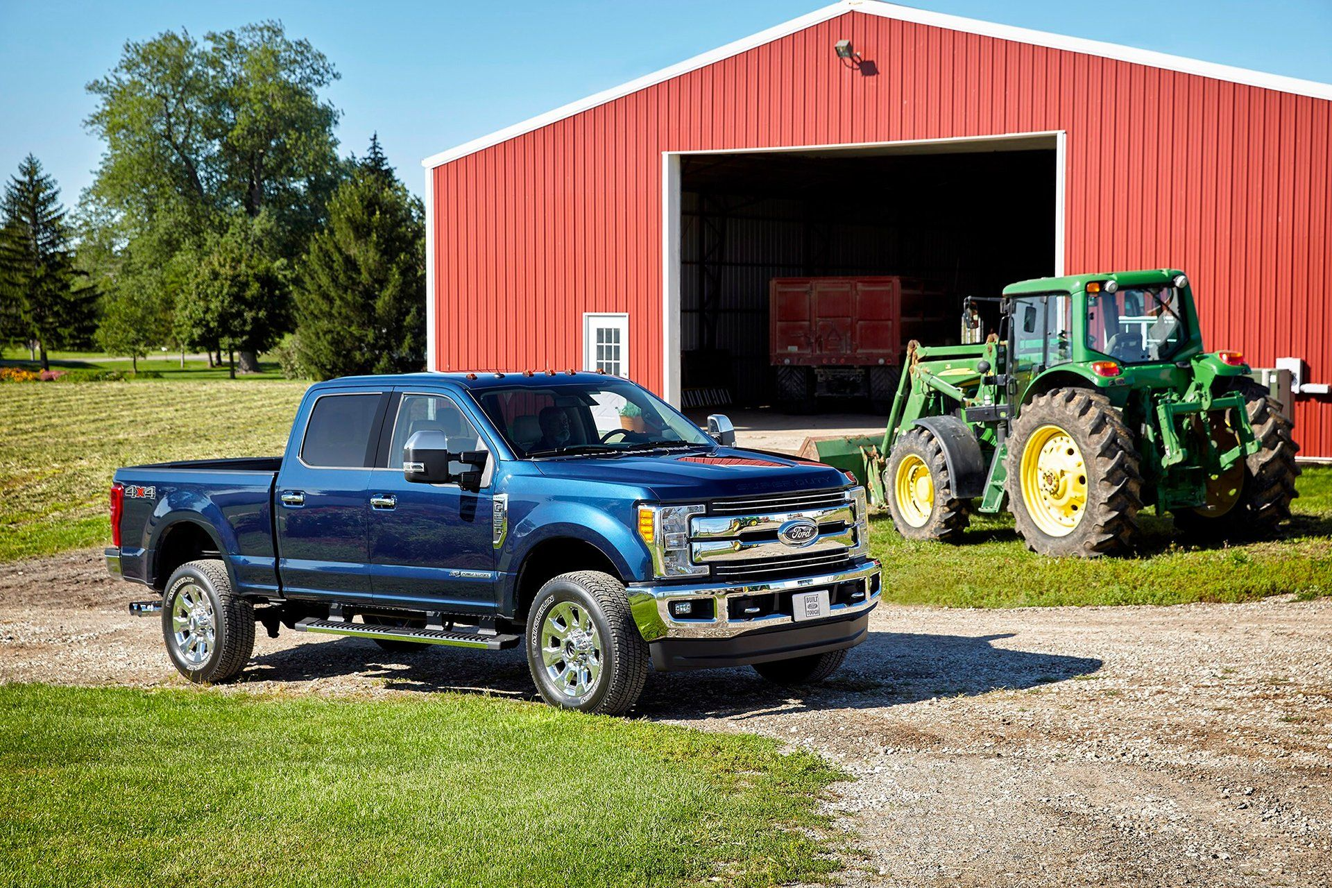 Blue pickup truck on a gravel drive, next to a green tractor and red barn. Sunny day.