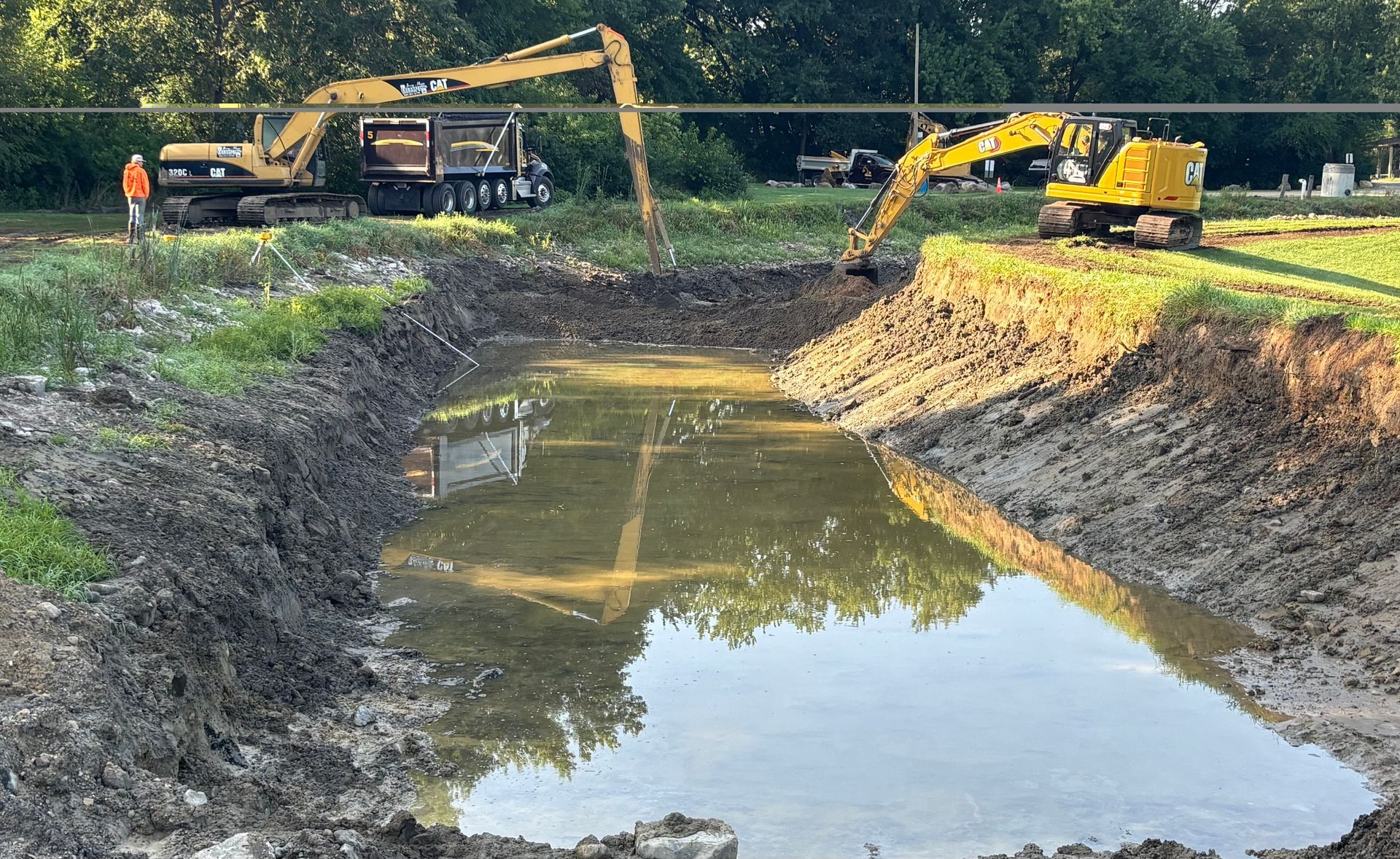 Two excavators beside a muddy pond at a construction site, with trees in the background.