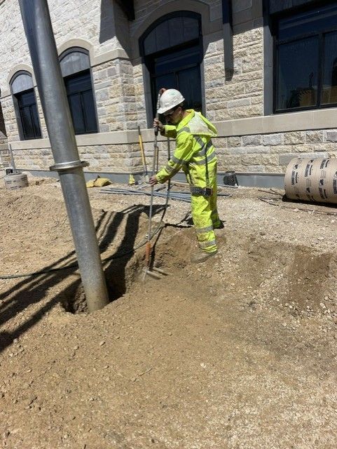 Construction worker in yellow safety gear digs near a metal pole, next to a stone building.