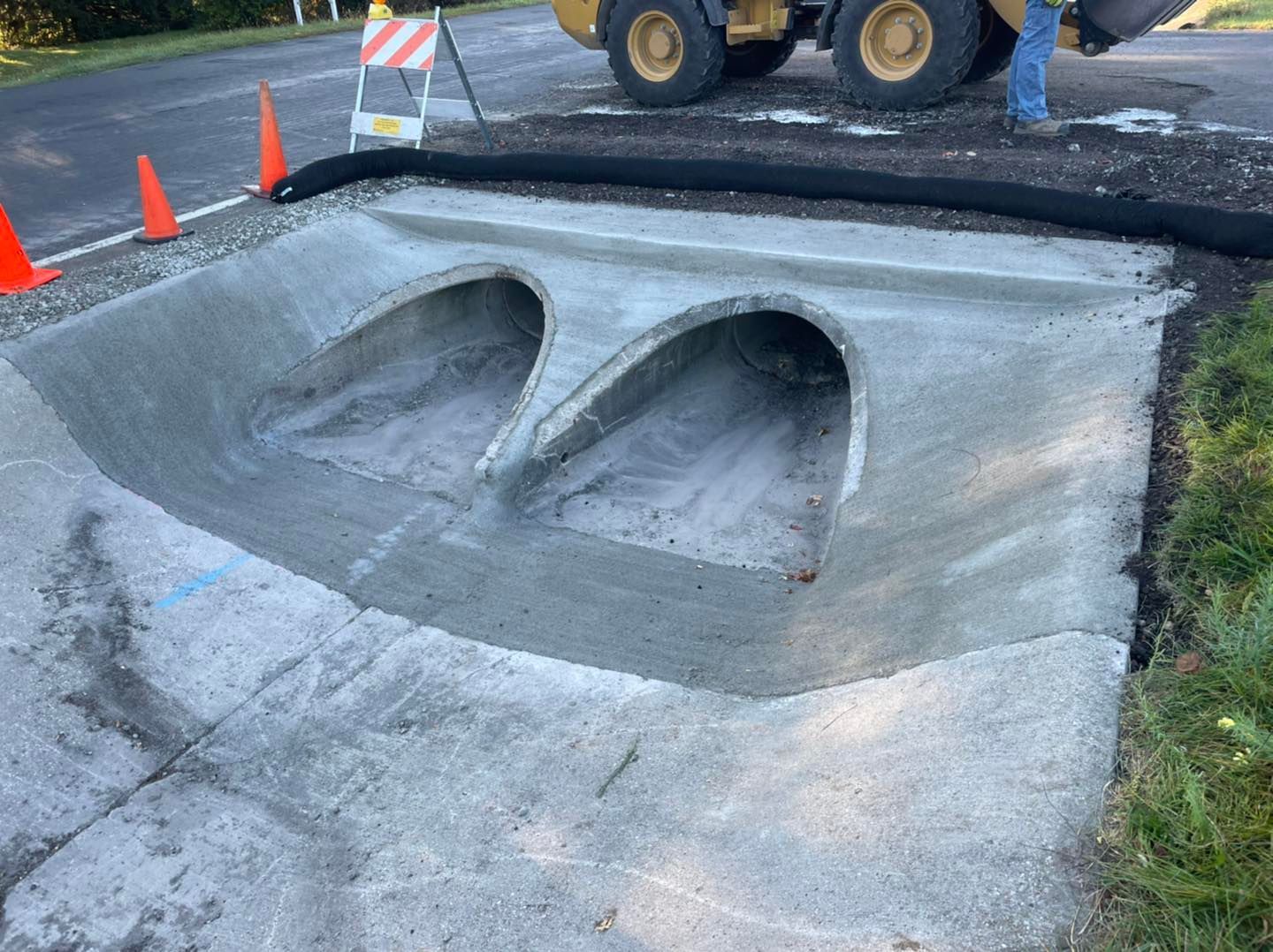 Concrete drainage structure with two arched openings, under construction near a road, with safety cones.