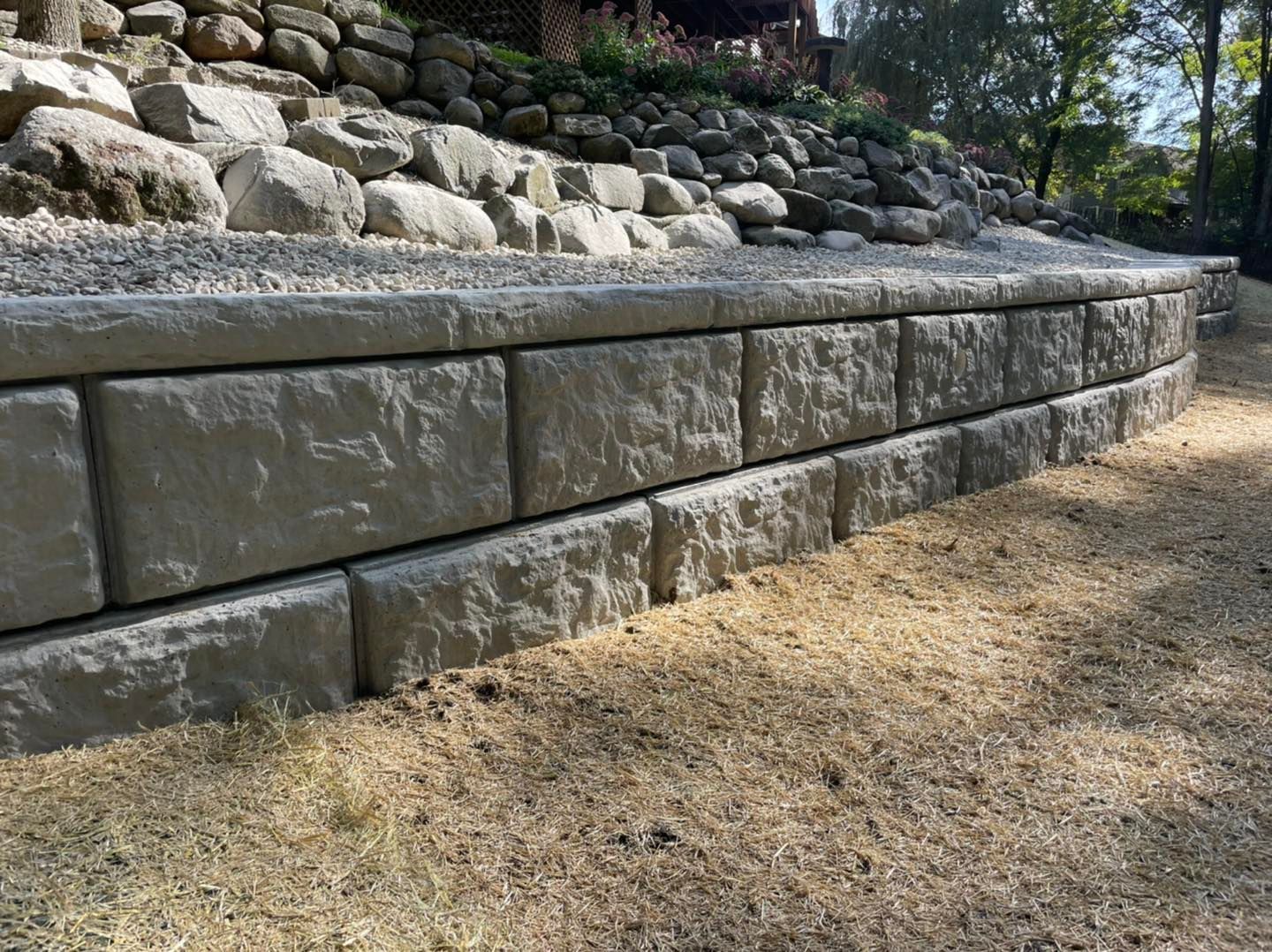 Retaining wall made of rectangular gray blocks, holding back a sloped area with rocks and gravel.