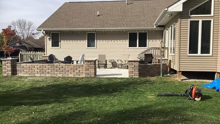 Backyard with a brick patio, beige house, and green lawn. A leaf blower is on the grass.