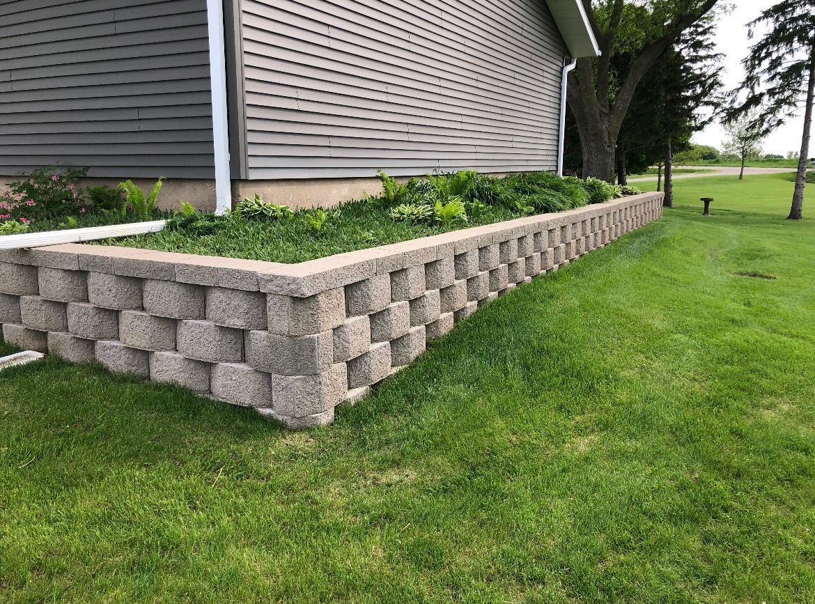 Stone retaining wall with a raised garden bed against a gray house; green grass surrounds.