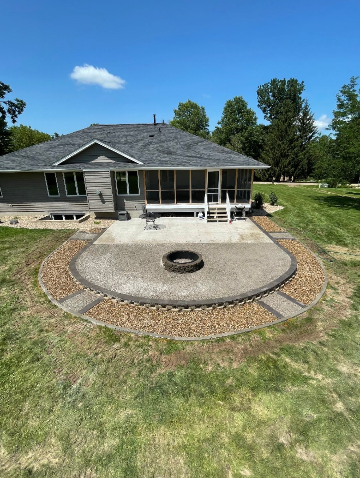 Backyard patio with fire pit, bordered by stone and rock, adjacent to a gray house with a screened porch.