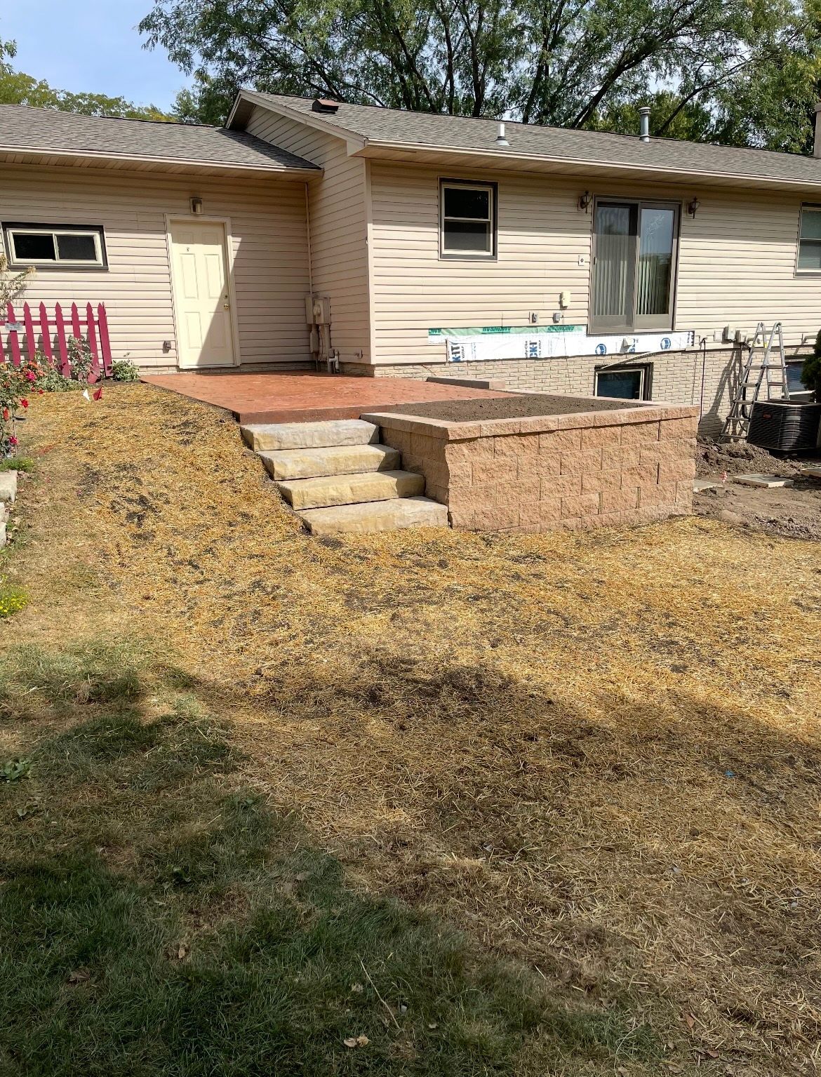 Backyard with a raised patio, steps, and a retaining wall. Tan siding, dry grass, and a sunny sky.