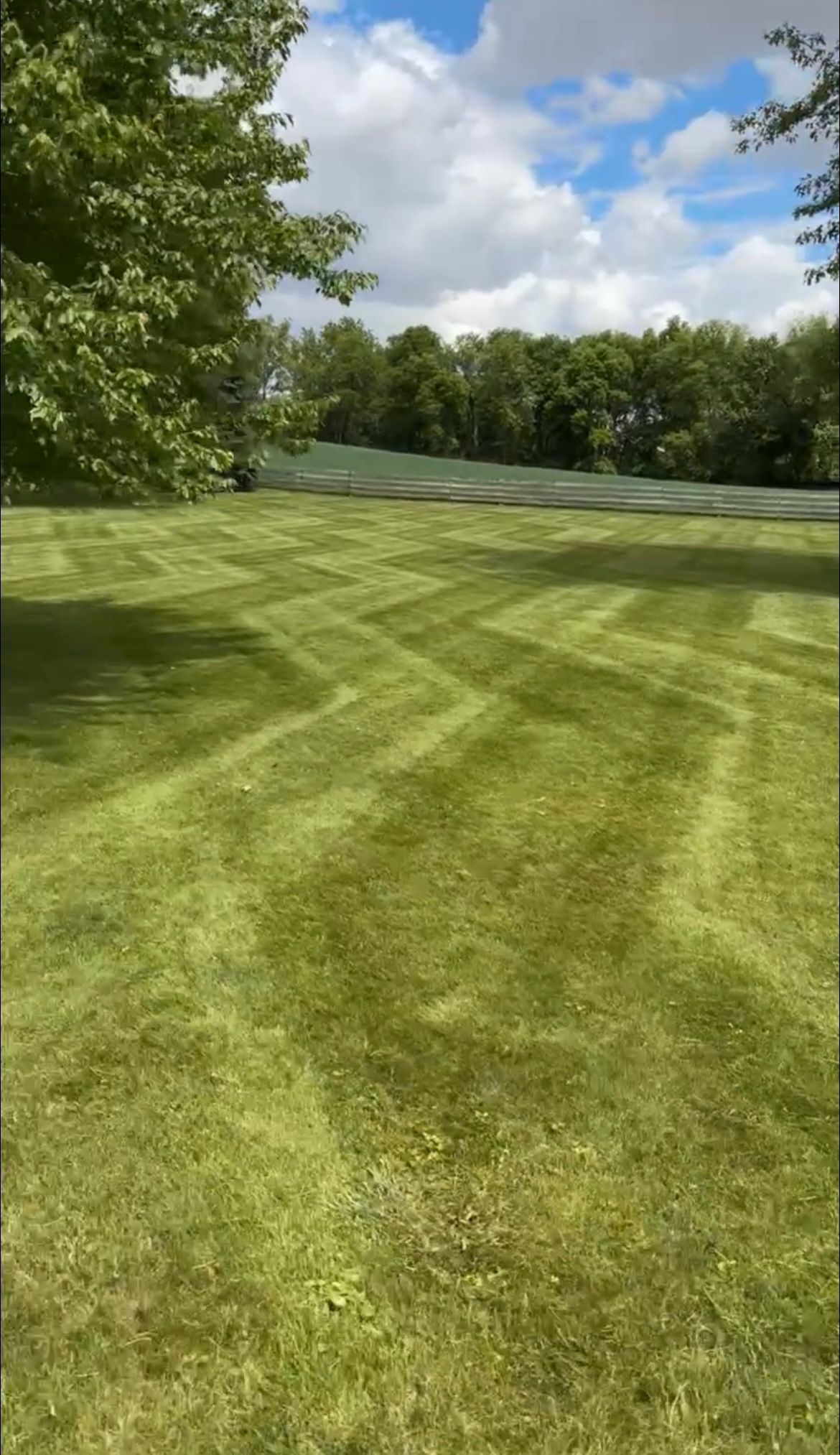 Lawn with freshly cut grass in a chevron pattern, green trees and blue sky with clouds in the background.