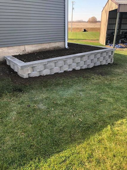 Gray retaining wall with dark soil against a gray house, next to a grassy lawn.