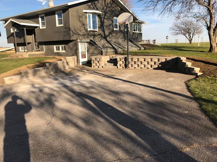 Asphalt driveway leading to a gray house with a basketball hoop. A concrete retaining wall borders the driveway.