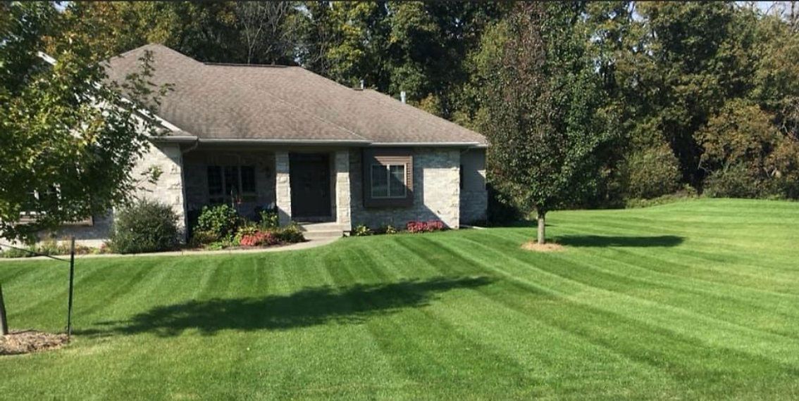 A well-manicured lawn with striped grass leads to a stone house with a covered porch. Lush trees in the background.