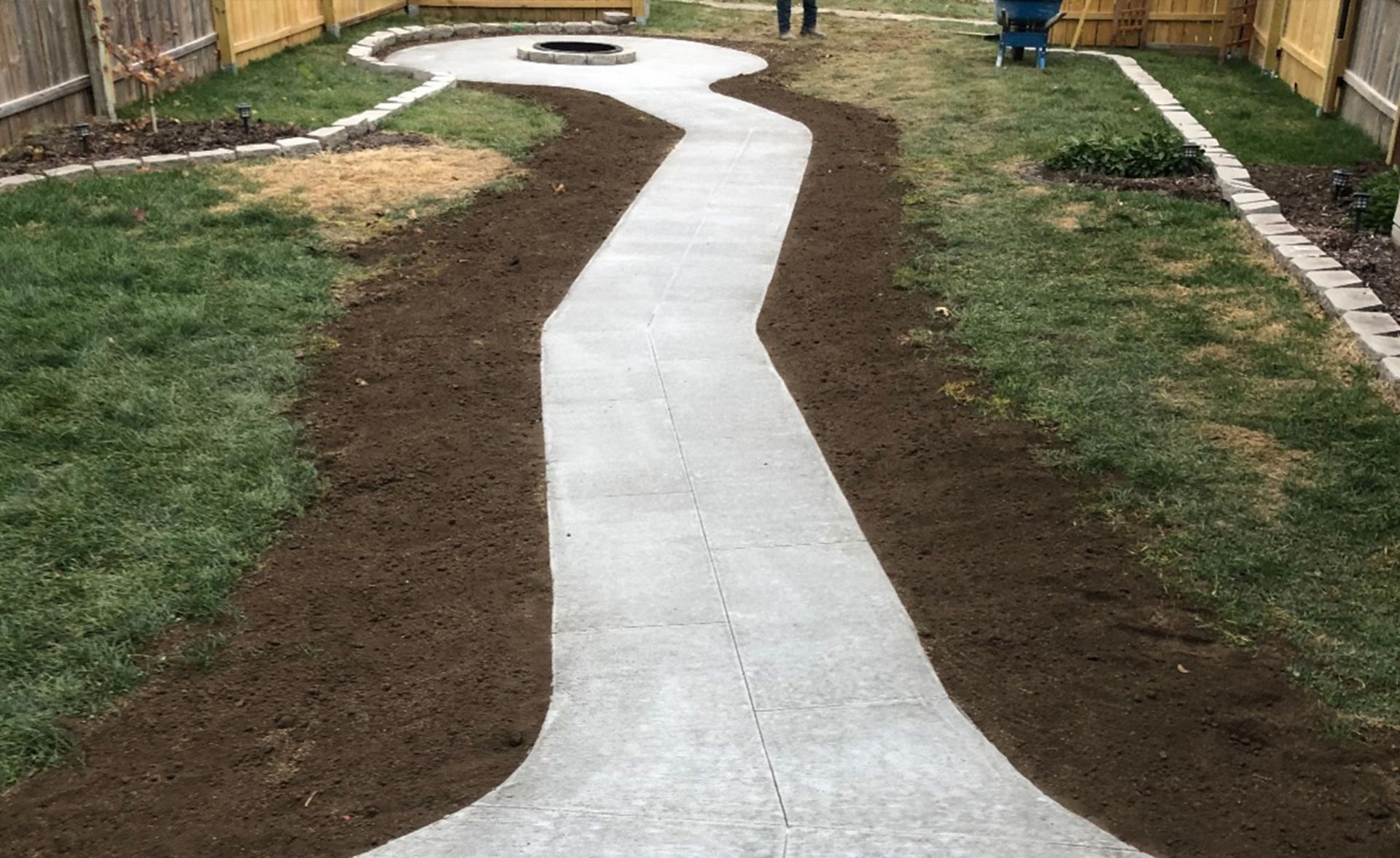 Concrete path in backyard, flanked by dirt beds and grass, leading to fire pit.