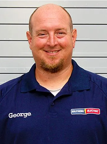 Smiling man in navy polo shirt with company logo, standing against a light gray wall.