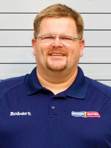Man in blue work shirt with name patch, smiling in front of a gray striped wall
