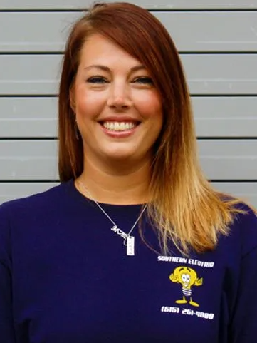 Smiling woman in a purple shirt standing against a gray wall, wearing a necklace and name badge