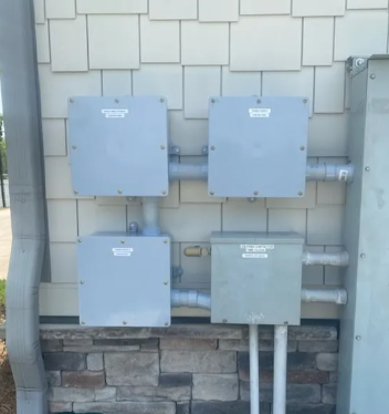 Four gray electrical utility boxes mounted on an exterior wall above stonework and pipes.
