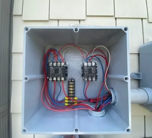 Open outdoor electrical junction box with wiring, relays, and terminal blocks mounted on a house exterior.