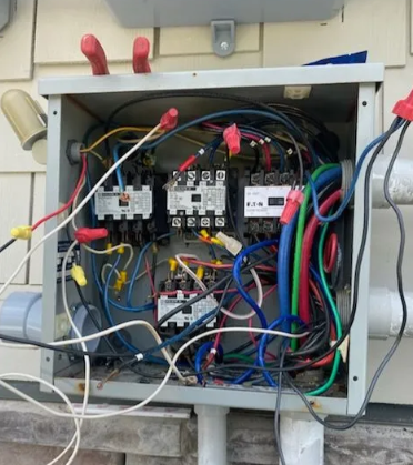 Open electrical control panel with tangled multicolored wiring and circuit components mounted on a wall.