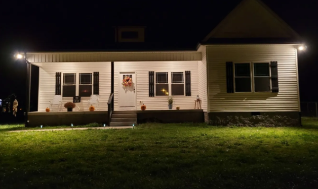 Single-story house at night with porch lights on, white siding, dark shutters, and a front lawn