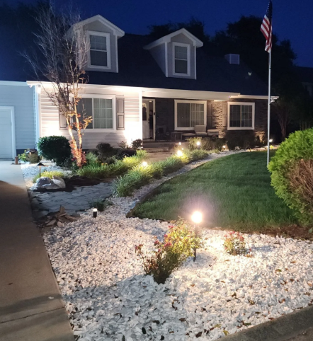 Front yard at night with white gravel landscaping, lit path lights, and a dark house with a porch and dormer windows